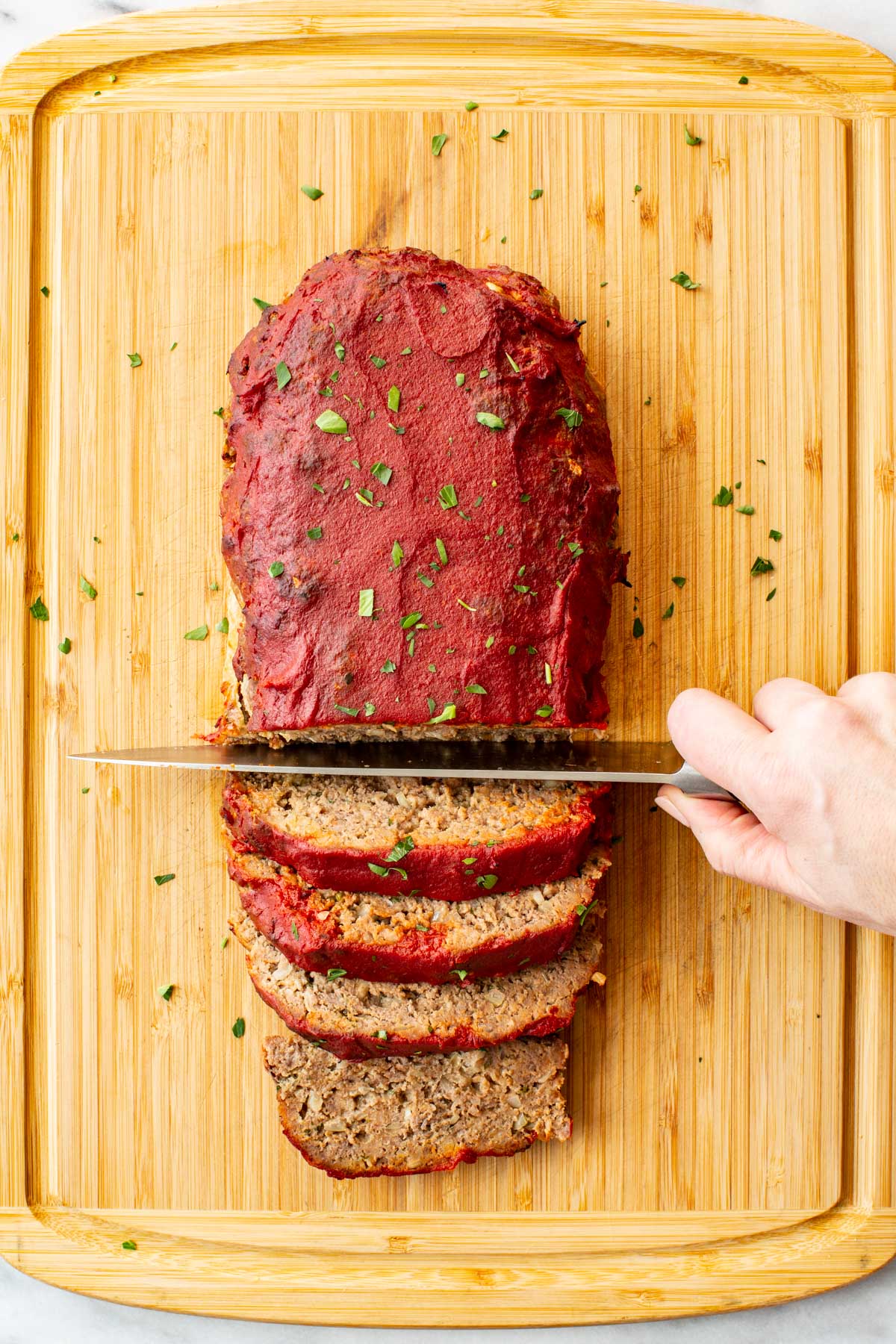 A hand slicing an Italian meatloaf on a wooden cutting board, with several thick slices already cut, showing the juicy interior and vibrant red tomato paste glaze, garnished with fresh chopped parsley.