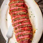 Overhead view of a fully sliced Italian meatloaf arranged on an oval ceramic platter, each slice showing a rich tomato glaze topping and hearty meat interior, scattered with fresh parsley, with a vintage serving knife on the side.