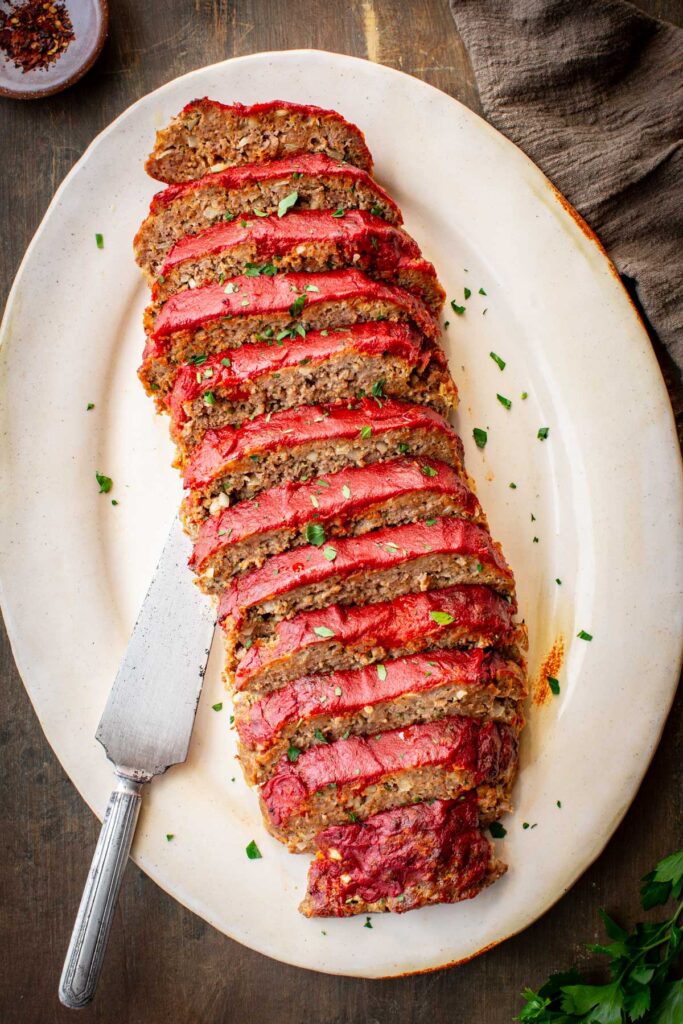 Overhead view of a fully sliced Italian meatloaf arranged on an oval ceramic platter, each slice showing a rich tomato glaze topping and hearty meat interior, scattered with fresh parsley, with a vintage serving knife on the side.