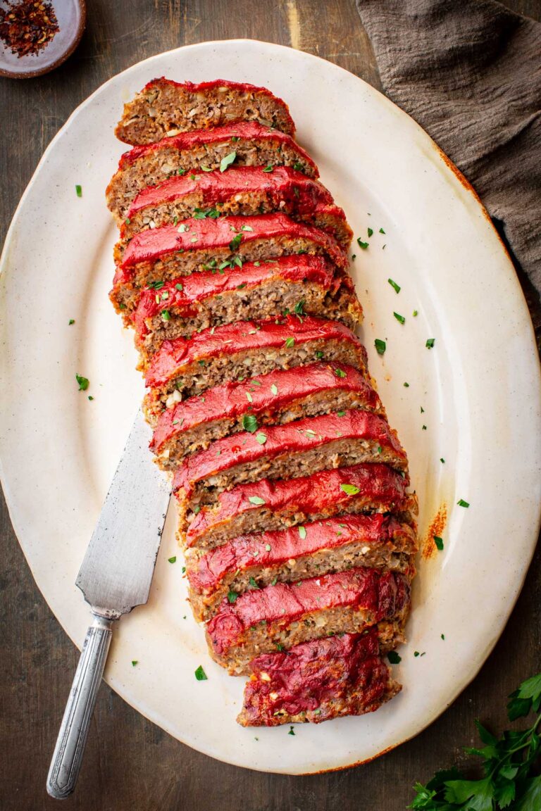 Overhead view of a fully sliced Italian meatloaf arranged on an oval ceramic platter, each slice showing a rich tomato glaze topping and hearty meat interior, scattered with fresh parsley, with a vintage serving knife on the side.