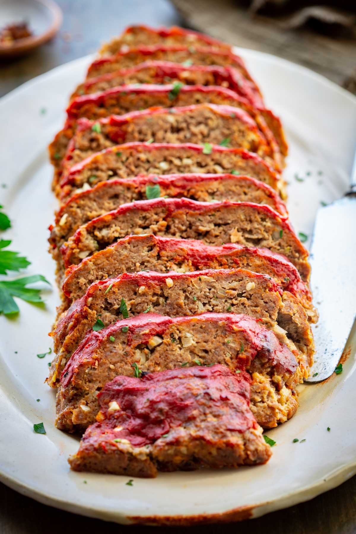 Close-up angled shot of Italian meatloaf slices fanned out on a ceramic platter, highlighting the caramelized tomato glaze, tender crumb texture, and fresh herb garnish.