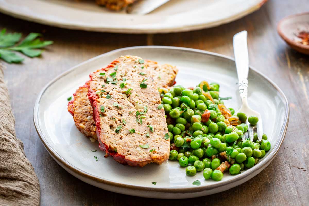 A serving plate with two slices of Italian meatloaf topped with red tomato glaze and chopped parsley, alongside a side of sautéed green peas, with a silver fork resting on the plate.
