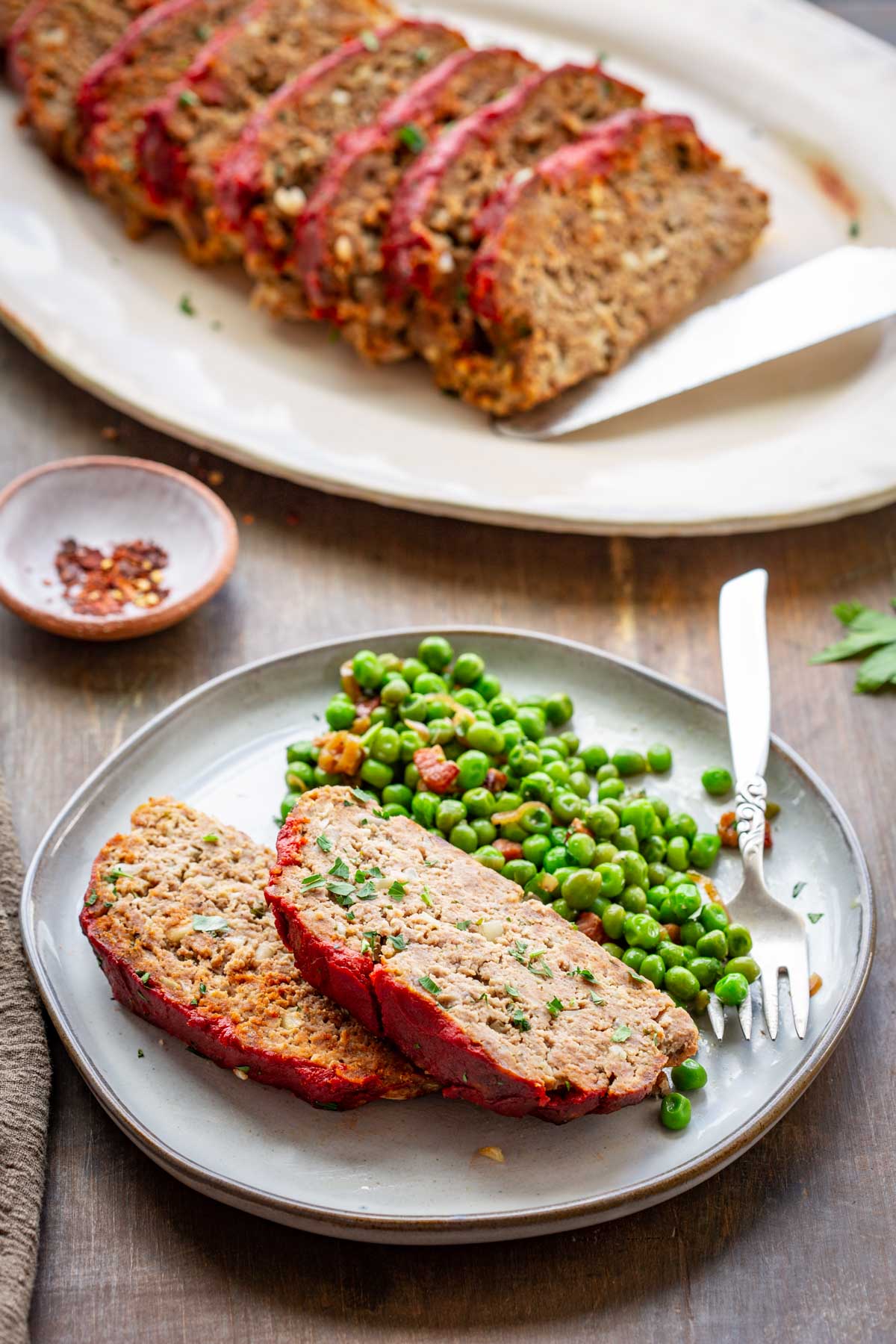 A rustic table setting with a dinner plate holding two slices of Italian meatloaf and a side of green peas, with the full sliced meatloaf on a platter in the background and a small bowl of red pepper flakes nearby.
