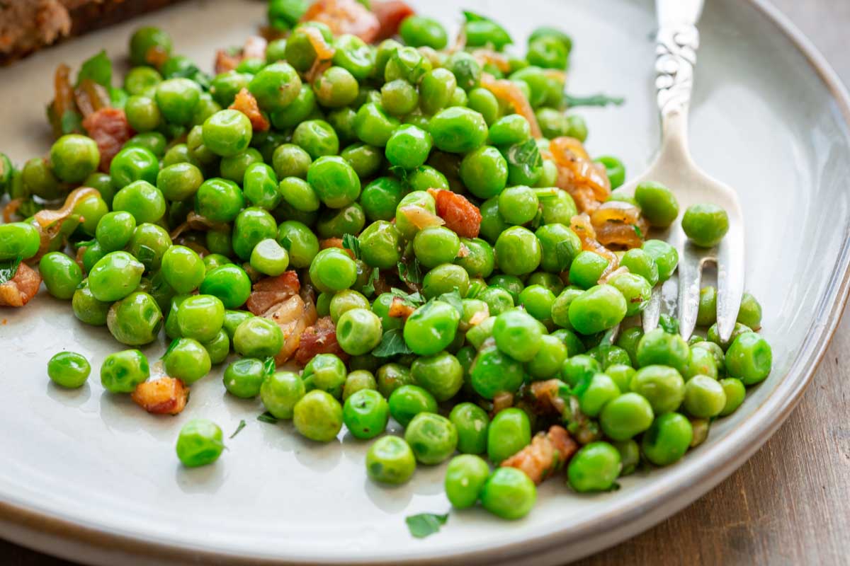 A serving of peas and pancetta with fresh parsley on a gray plate with a fork, shown as a side dish alongside a main course.