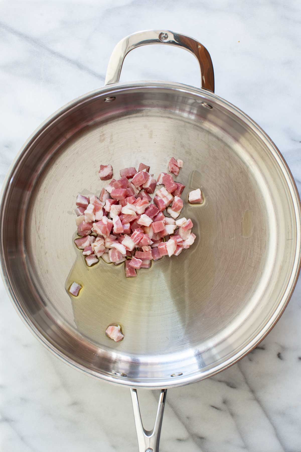 Raw diced pancetta in a stainless steel skillet with a small amount of olive oil, ready to cook.