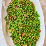 Peas with pancetta garnished with fresh chopped parsley, served on a large white oval platter with a serving spoon.