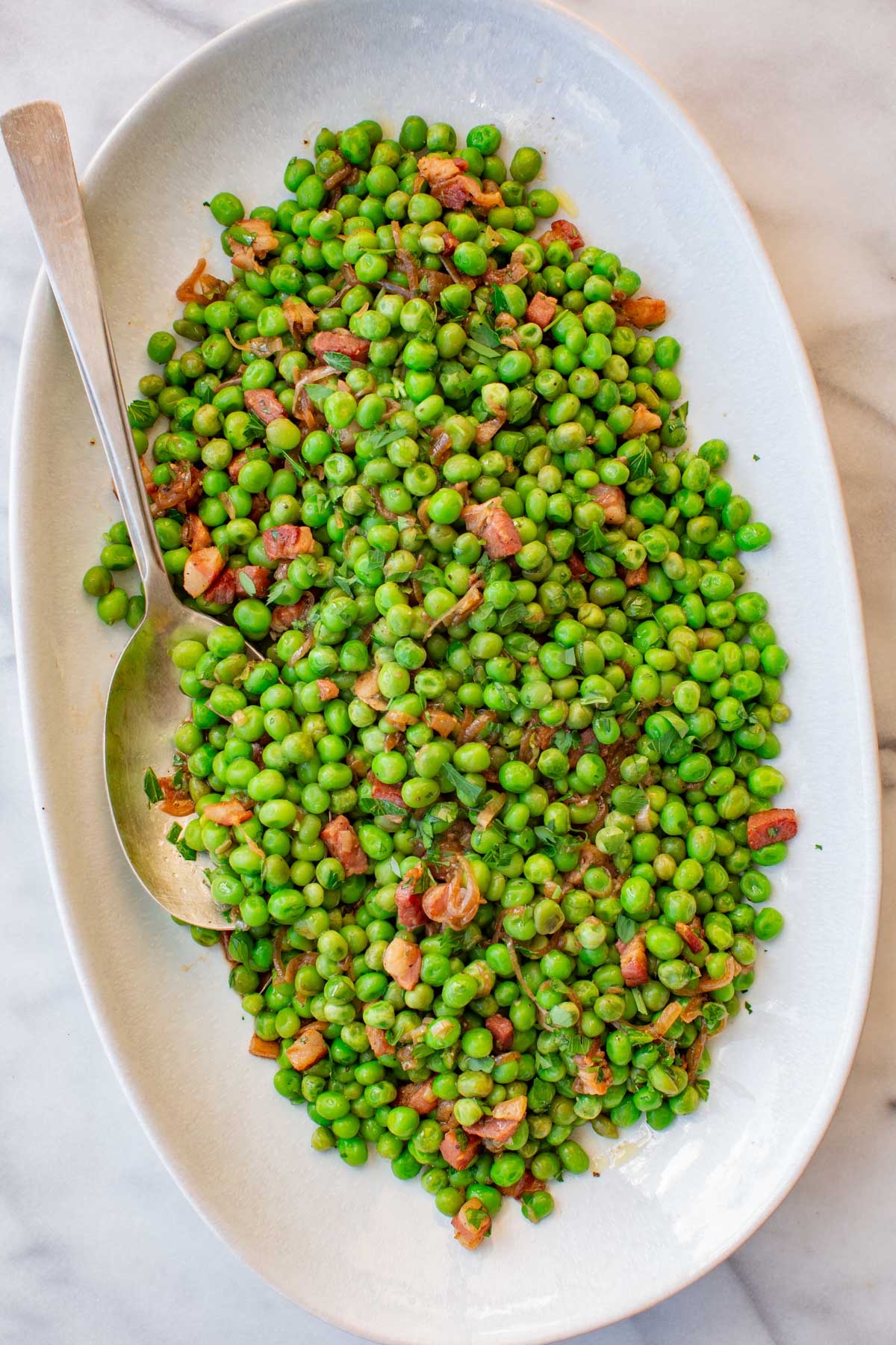 Peas with pancetta garnished with fresh chopped parsley, served on a large white oval platter with a serving spoon.