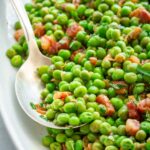 Close-up of peas and pancetta with fresh herbs on a white platter, with a serving spoon scooping a portion.
