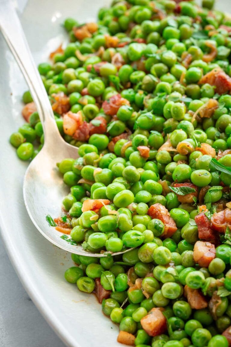 Close-up of peas and pancetta with fresh herbs on a white platter, with a serving spoon scooping a portion.