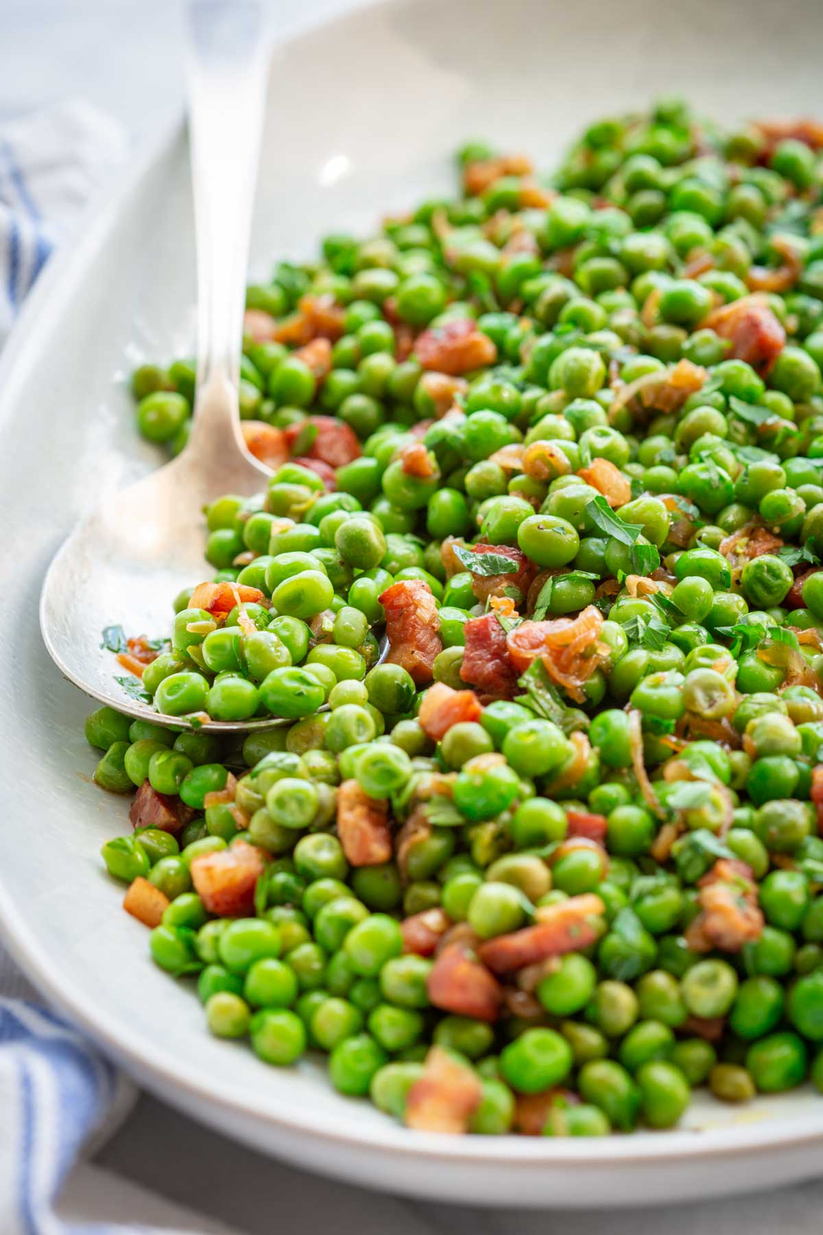 Angled close-up of peas and pancetta with chopped parsley on a white oval platter with a serving spoon.