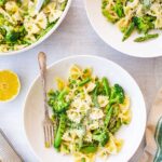 Overhead view of two white bowls and a serving pan of pasta primavera on a light gray surface, featuring farfalle (bow-tie pasta) tossed with broccoli, asparagus, snap peas, and green peas, garnished with grated parmesan, with a halved lemon and a block of parmesan cheese on the side.