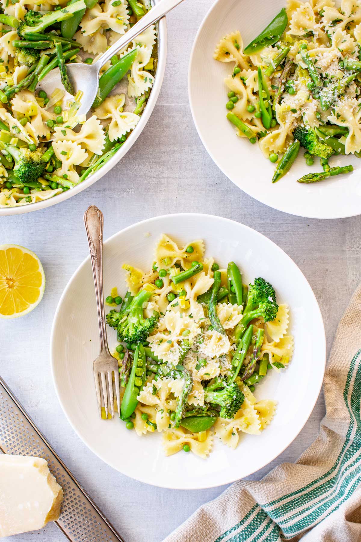 Overhead view of two white bowls and a serving pan of pasta primavera on a light gray surface, featuring farfalle (bow-tie pasta) tossed with broccoli, asparagus, snap peas, and green peas, garnished with grated parmesan, with a halved lemon and a block of parmesan cheese on the side.