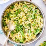 Overhead shot of a large stainless steel skillet filled with pasta primavera - farfalle tossed with broccoli florets, snap peas, asparagus, peas, and sautéed leeks - with a serving spoon resting inside, surrounded by a lemon half, parmesan block, and striped linen napkin.