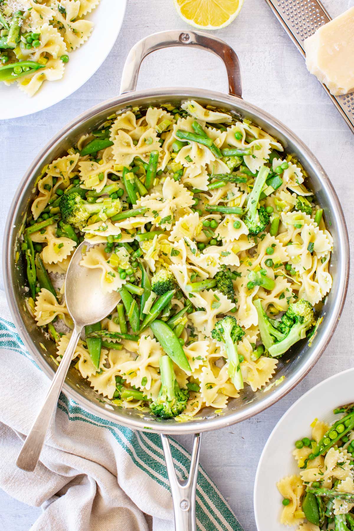 Overhead shot of a large stainless steel skillet filled with pasta primavera - farfalle tossed with broccoli florets, snap peas, asparagus, peas, and sautéed leeks - with a serving spoon resting inside, surrounded by a lemon half, parmesan block, and striped linen napkin.