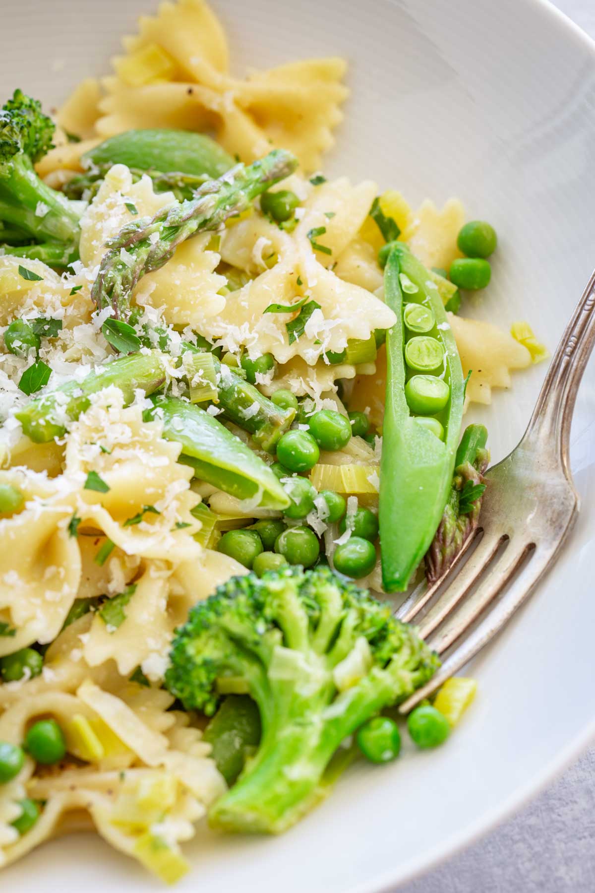 Close-up of a white bowl of pasta primavera, showcasing farfalle pasta with vibrant green broccoli florets, snap peas, asparagus spears, and green peas, generously topped with freshly grated parmesan and chopped herbs, with a vintage fork resting in the bowl.