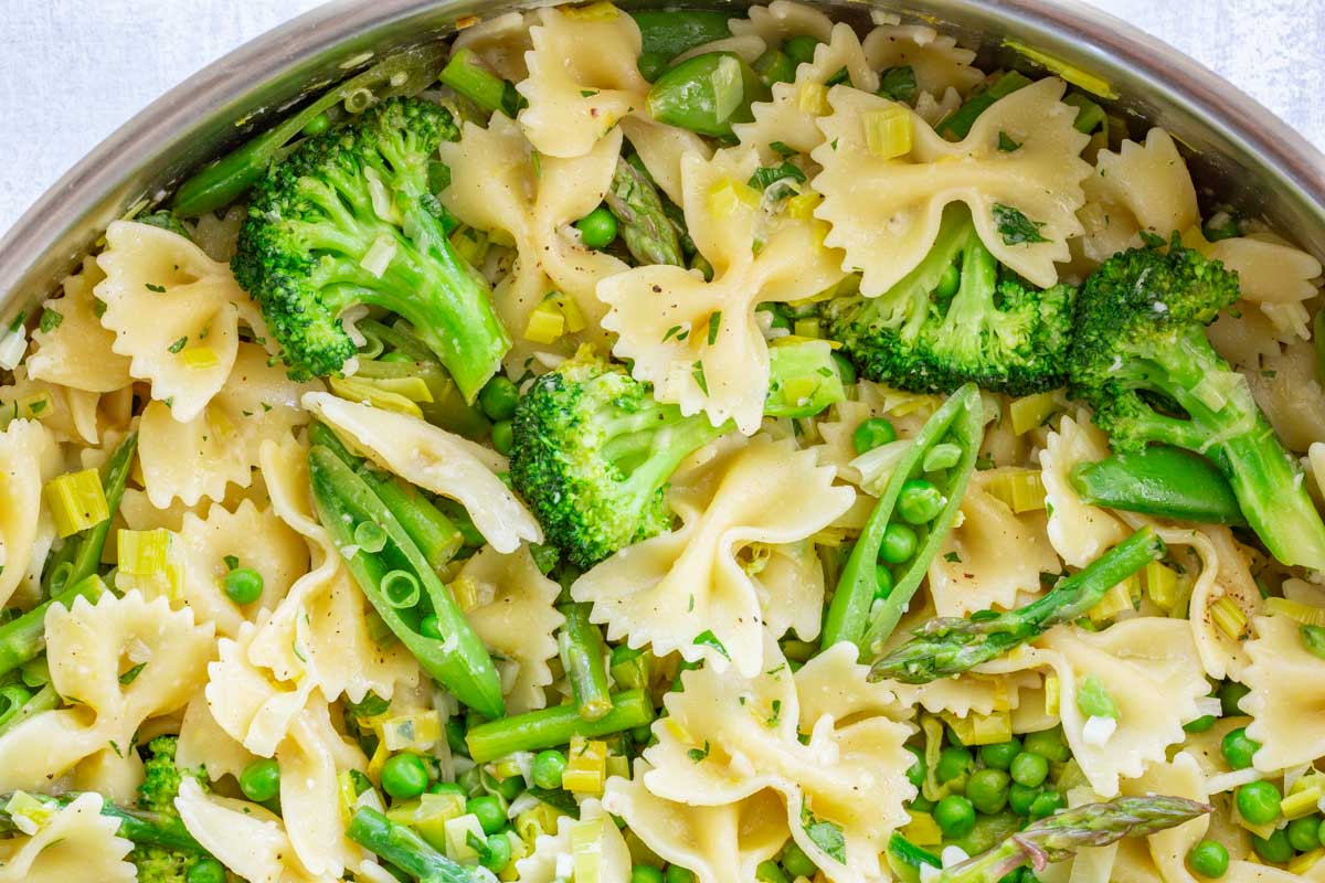 Close-up overhead view inside a stainless steel skillet packed with pasta primavera, highlighting the abundance of green vegetables - broccoli florets, asparagus pieces, snap peas, and peas - mixed with farfalle pasta and sautéed leeks.