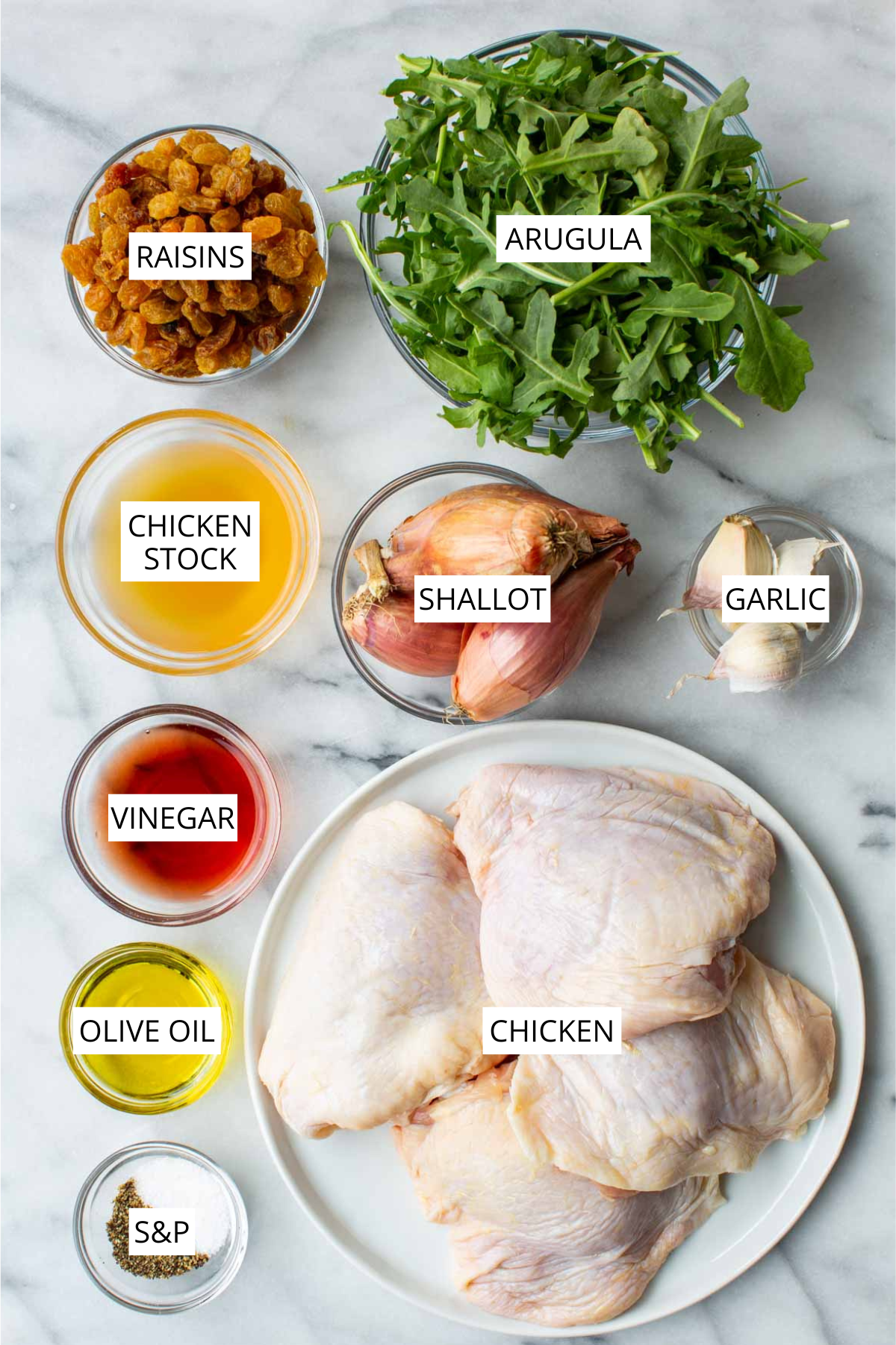 Overhead shot of chicken agrodolce ingredients arranged on a white marble surface: a plate of raw bone-in, skin-on chicken thighs, a bowl of golden raisins, a large bowl of fresh arugula, a bowl of chicken stock, a whole shallot, a small bowl of garlic cloves, a small bowl of red wine vinegar, a small bowl of olive oil, and a small bowl of salt and pepper.