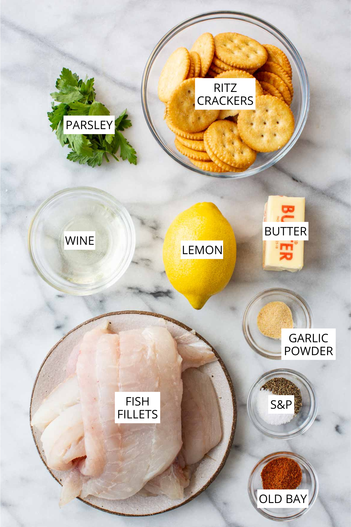 Flat lay of all recipe ingredients on a marble surface, labeled: Ritz crackers in a glass bowl, a bunch of parsley, a small bowl of white wine, a whole lemon, a stick of butter, a small bowl of garlic powder, two small bowls of salt and pepper, a plate of raw fish fillets, and a small bowl of Old Bay seasoning.