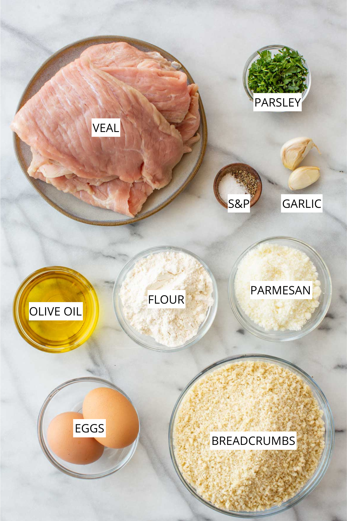 Overhead flat lay of veal Milanese ingredients on a marble surface, labeled: raw veal cutlets on a plate, fresh parsley in a small bowl, salt and pepper in a small wooden bowl, two garlic cloves, a bowl of olive oil, a bowl of all-purpose flour, a bowl of grated parmesan, two eggs in a bowl, and a large bowl of breadcrumbs.