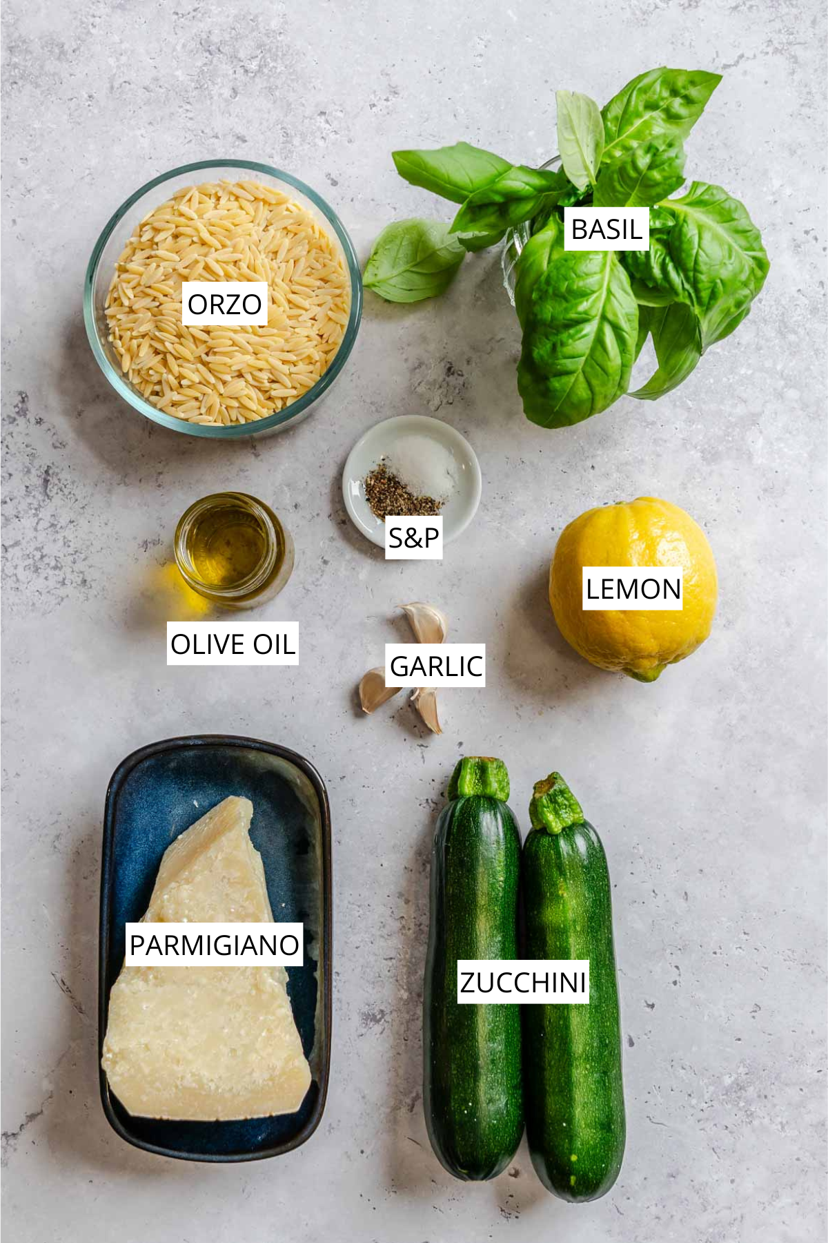 Flat lay of labeled ingredients for zucchini orzo on a light gray surface: a glass bowl of orzo, a bunch of fresh basil, a small dish of salt and pepper, a lemon, two garlic cloves, a jar of olive oil, a wedge of Parmigiano on a dark tray, and two whole zucchini.