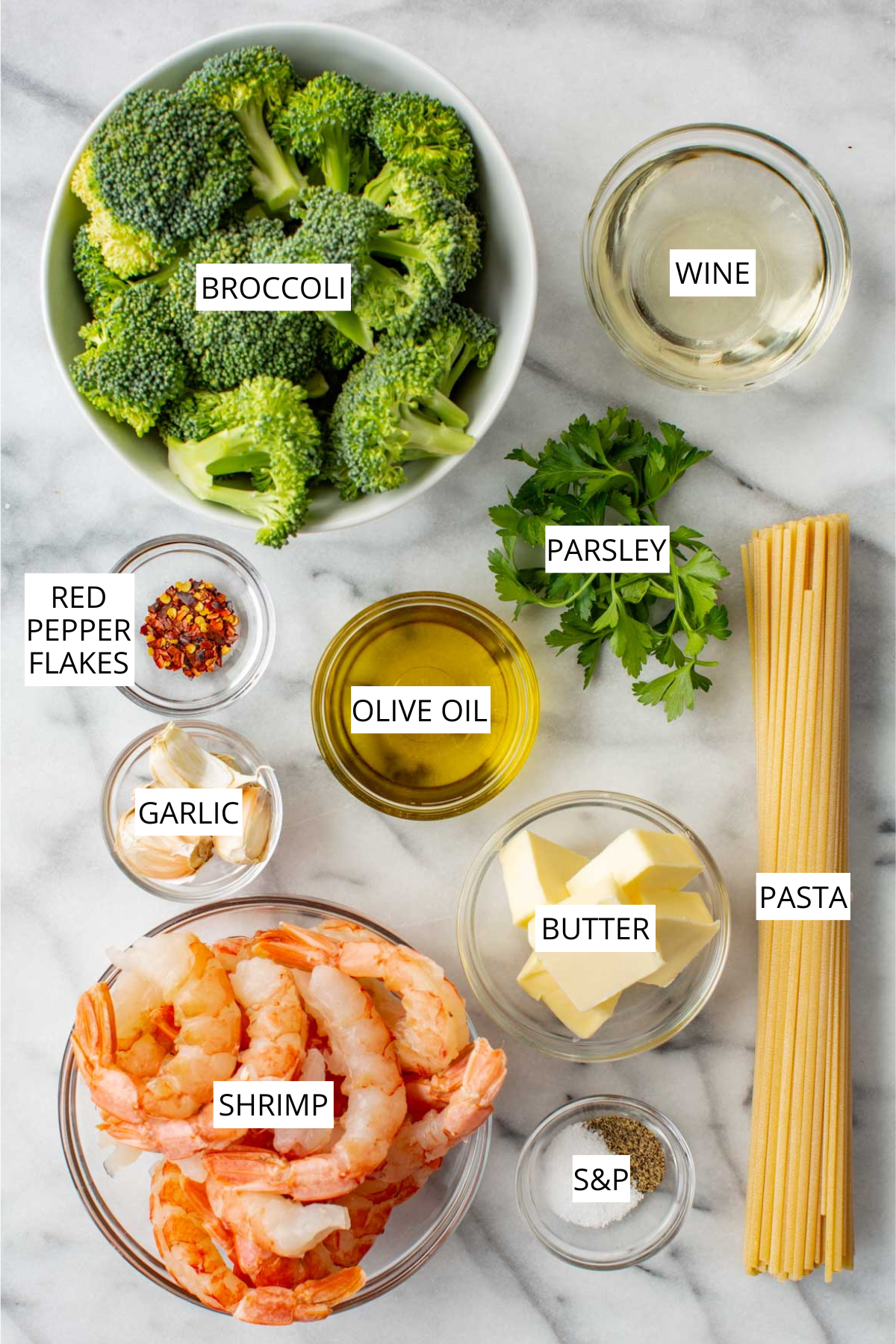 Flat lay of shrimp and broccoli pasta ingredients on a marble surface, labeled: broccoli, white wine, red pepper flakes, parsley, olive oil, garlic, pasta, butter, shrimp, and salt and pepper.