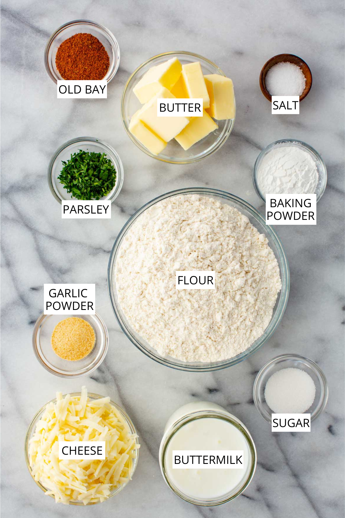 Overhead flat lay of cheddar bay biscuit ingredients on a marble surface, labeled: Old Bay seasoning, butter, salt, parsley, baking powder, garlic powder, flour, cheese, buttermilk, and sugar.
