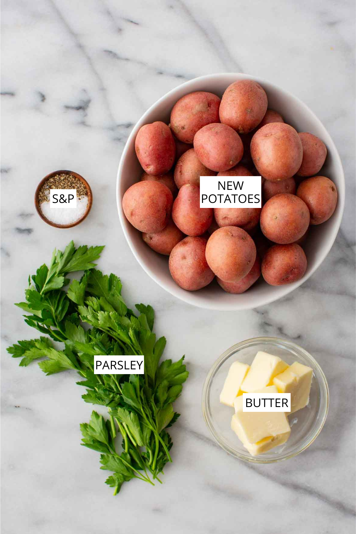 Flat lay of four ingredients on a white marble surface: a white bowl of whole new potatoes, a bunch of fresh flat-leaf parsley, a glass bowl with three pats of butter, and a small wooden bowl with salt and pepper, each labeled with its name.