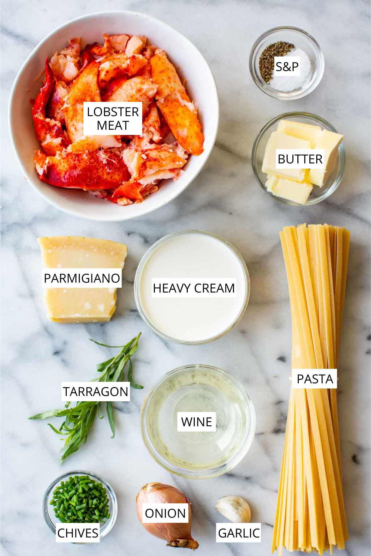Flat lay of all ingredients for lobster pasta arranged on a marble surface and labeled: a bowl of raw lobster meat, small bowls of salt and pepper, butter cubes, a wedge of Parmigiano, a glass of heavy cream, a bundle of tarragon, a glass of white wine, dry linguine, chopped chives, a shallot, and a garlic clove.