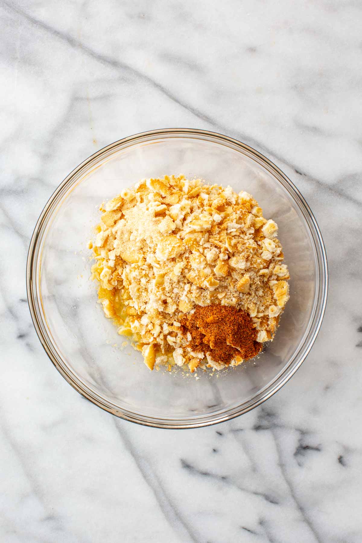 A glass bowl on a marble surface filled with crushed Ritz crackers, garlic powder, and a mound of Old Bay seasoning, before mixing.