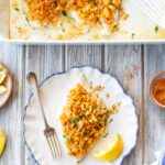 Split overhead scene: the white baking dish with the full batch of Ritz-topped fish above, and a single serving plated on a blue-rimmed dish below, accompanied by a lemon wedge and a fork, with small bowls of parsley, Old Bay, and lemon nearby.