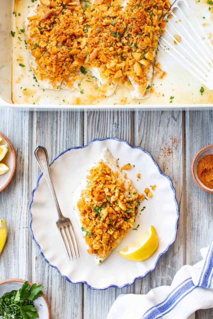 Split overhead scene: the white baking dish with the full batch of Ritz-topped fish above, and a single serving plated on a blue-rimmed dish below, accompanied by a lemon wedge and a fork, with small bowls of parsley, Old Bay, and lemon nearby.