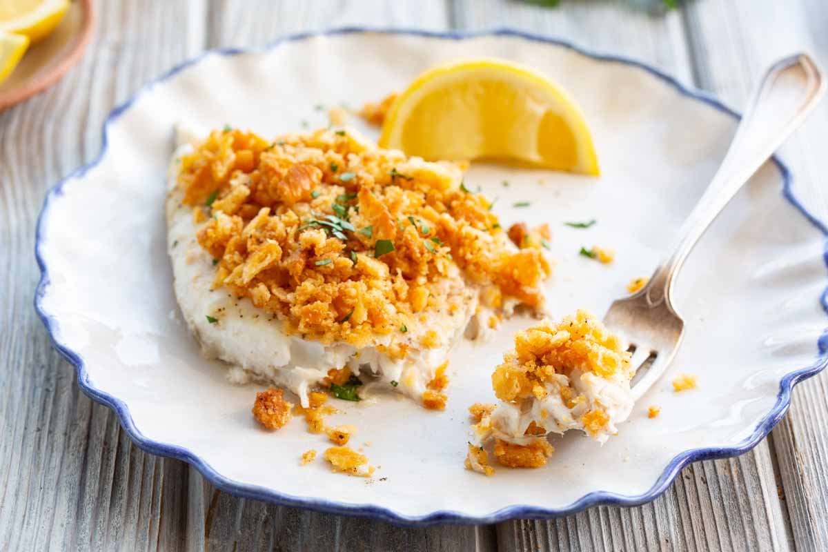 Close-up of a single baked fish fillet on a blue-rimmed white plate, the crispy Ritz cracker topping partially broken by a fork, served with two lemon wedges.