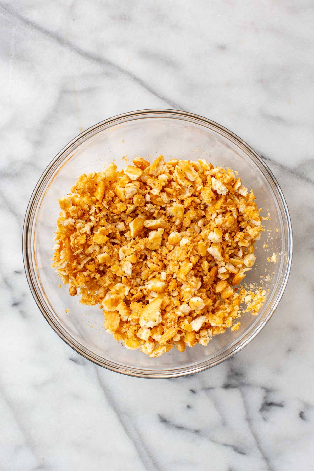 A glass bowl on a marble surface filled with the fully mixed Ritz cracker topping, evenly coated in golden-orange seasoning.