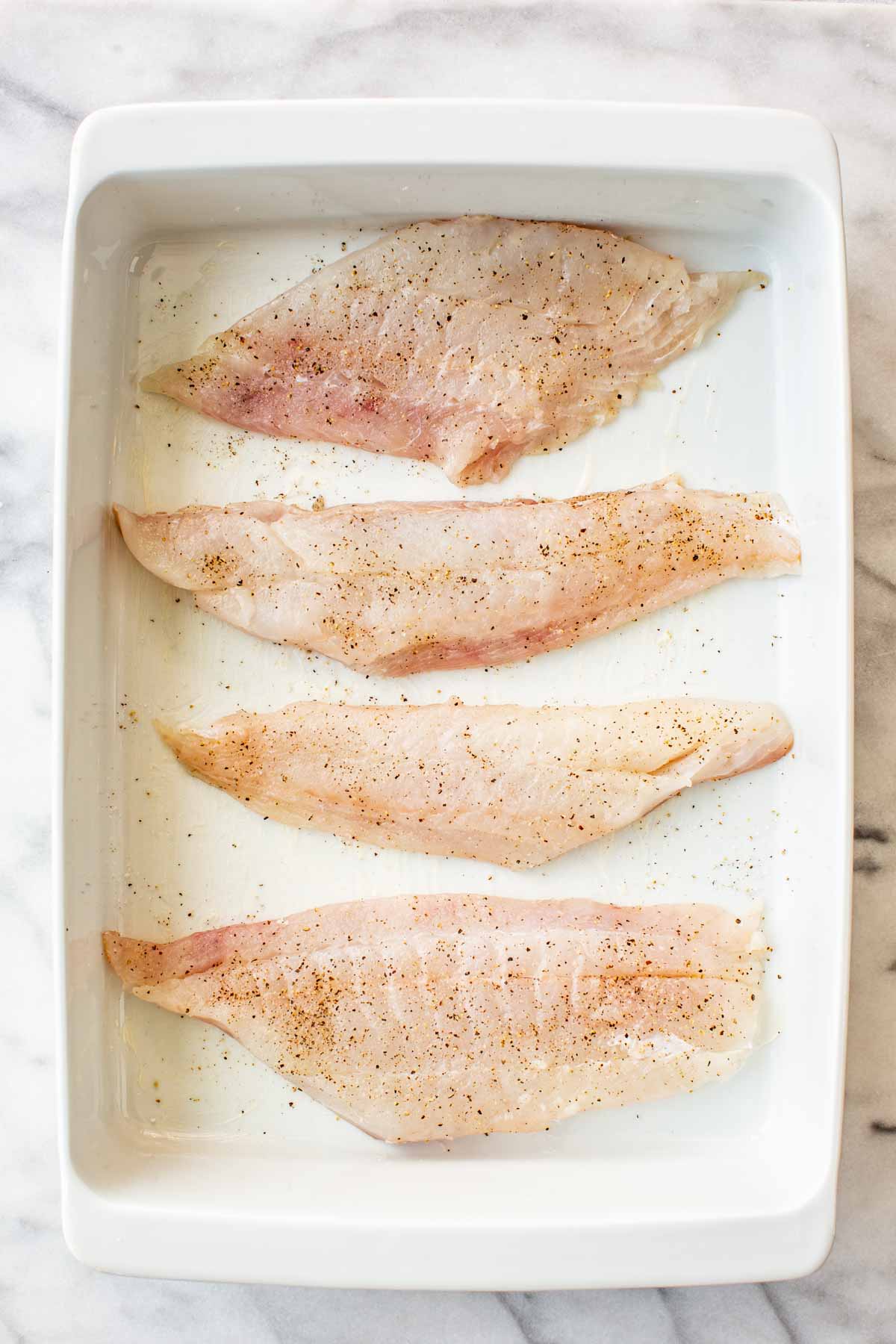 Four raw white fish fillets arranged in a white ceramic baking dish on a marble surface, seasoned with salt and pepper.