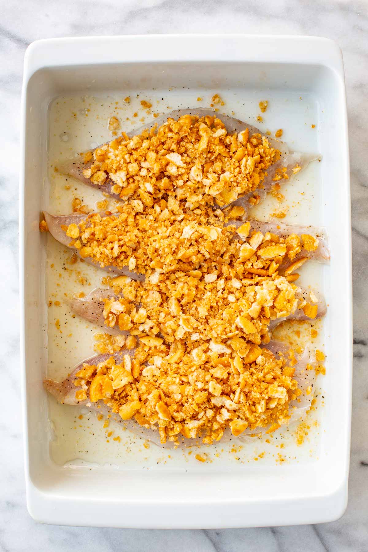 Four seasoned raw fish fillets in a white baking dish, each topped with a generous mound of the seasoned Ritz cracker mixture, ready to bake.