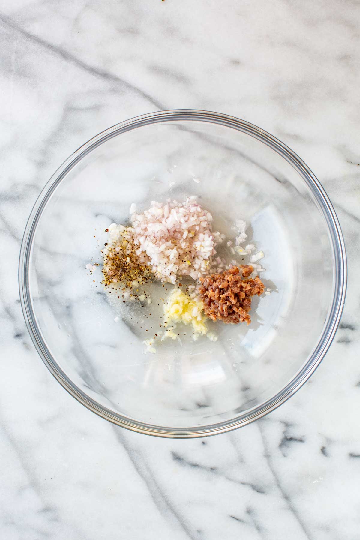 Overhead view of a large glass bowl containing minced shallot, mashed anchovy paste, minced garlic, salt, and black pepper - the start of a vinaigrette dressing.