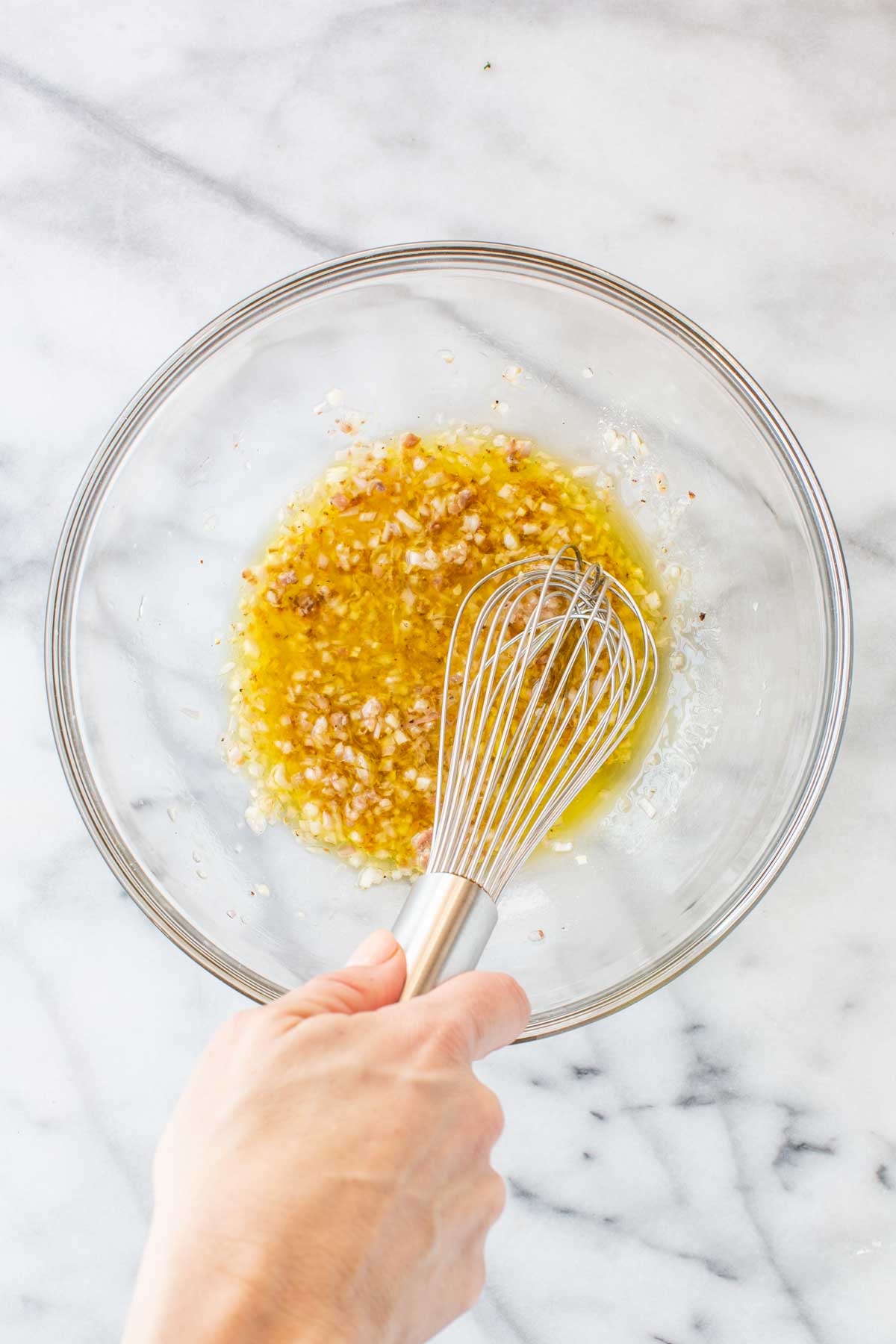 A hand whisking the anchovy vinaigrette in a glass bowl, with olive oil and vinegar emulsifying into a golden dressing with visible shallot and garlic.
