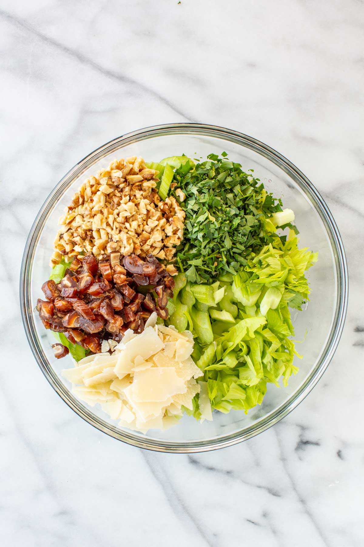 Overhead view of a glass bowl with the salad ingredients arranged in sections before tossing: chopped celery, chopped parsley, chopped walnuts, chopped dates, and shaved Parmesan.