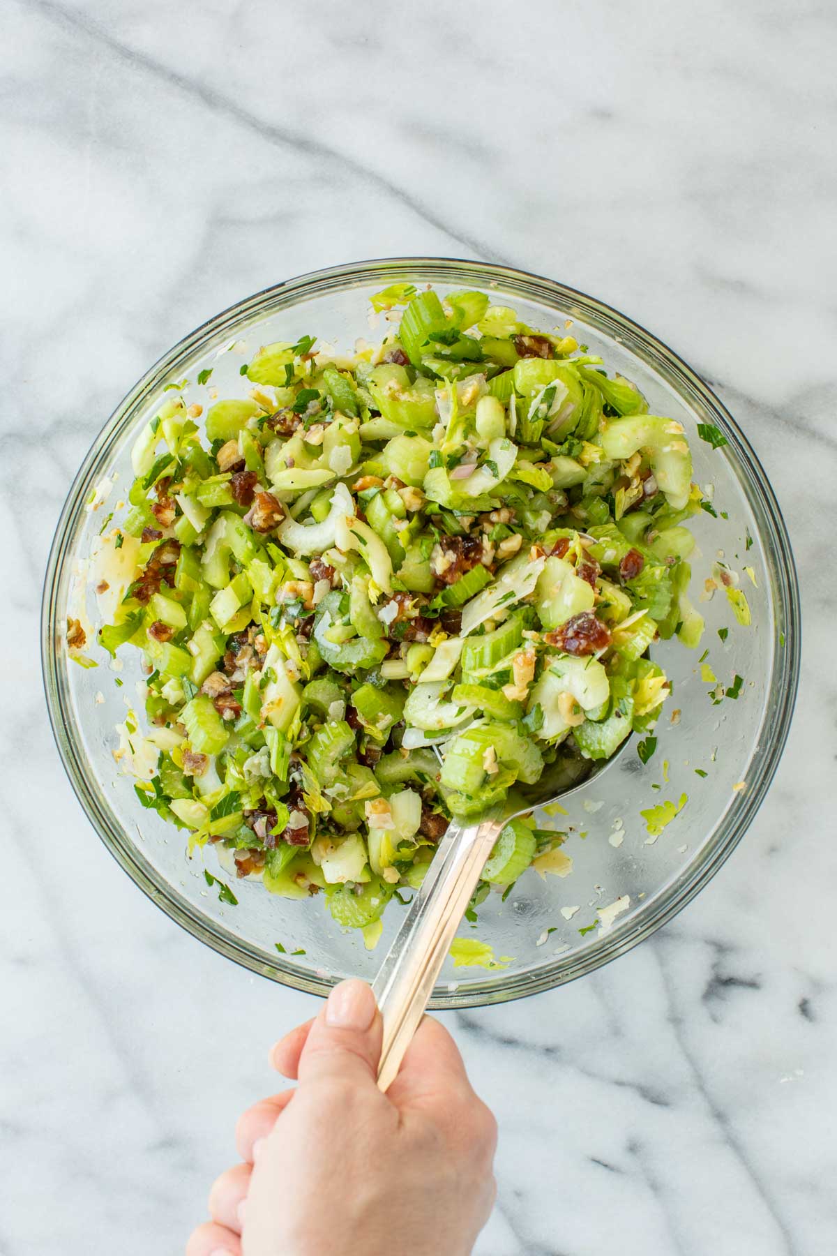 A hand tossing the celery salad in a glass bowl with a large spoon, all ingredients coated in the golden anchovy vinaigrette.