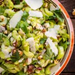 Close-up overhead view of the finished celery salad served in a striped ceramic bowl, topped with large shaved Parmesan curls, chopped walnuts, dates, and fresh parsley leaves.