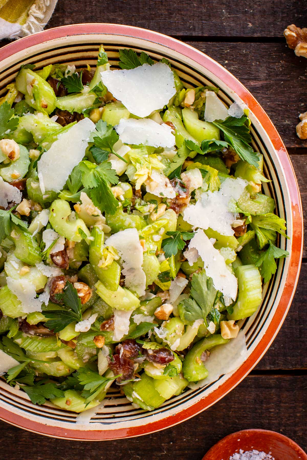 Close-up overhead view of the finished celery salad served in a striped ceramic bowl, topped with large shaved Parmesan curls, chopped walnuts, dates, and fresh parsley leaves.