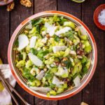 Full overhead shot of the celery salad in a striped ceramic bowl on a dark wood surface, surrounded by a tin of anchovies, a wedge of Parmesan, a dish of walnuts, flaky salt, and serving utensils.