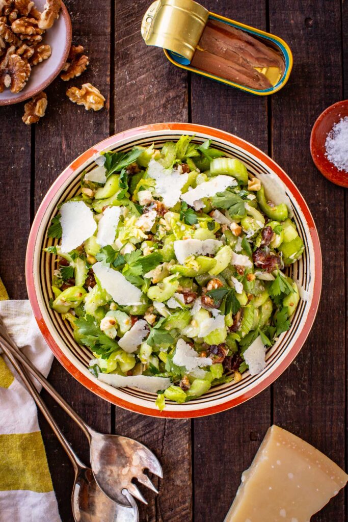 Full overhead shot of the celery salad in a striped ceramic bowl on a dark wood surface, surrounded by a tin of anchovies, a wedge of Parmesan, a dish of walnuts, flaky salt, and serving utensils.