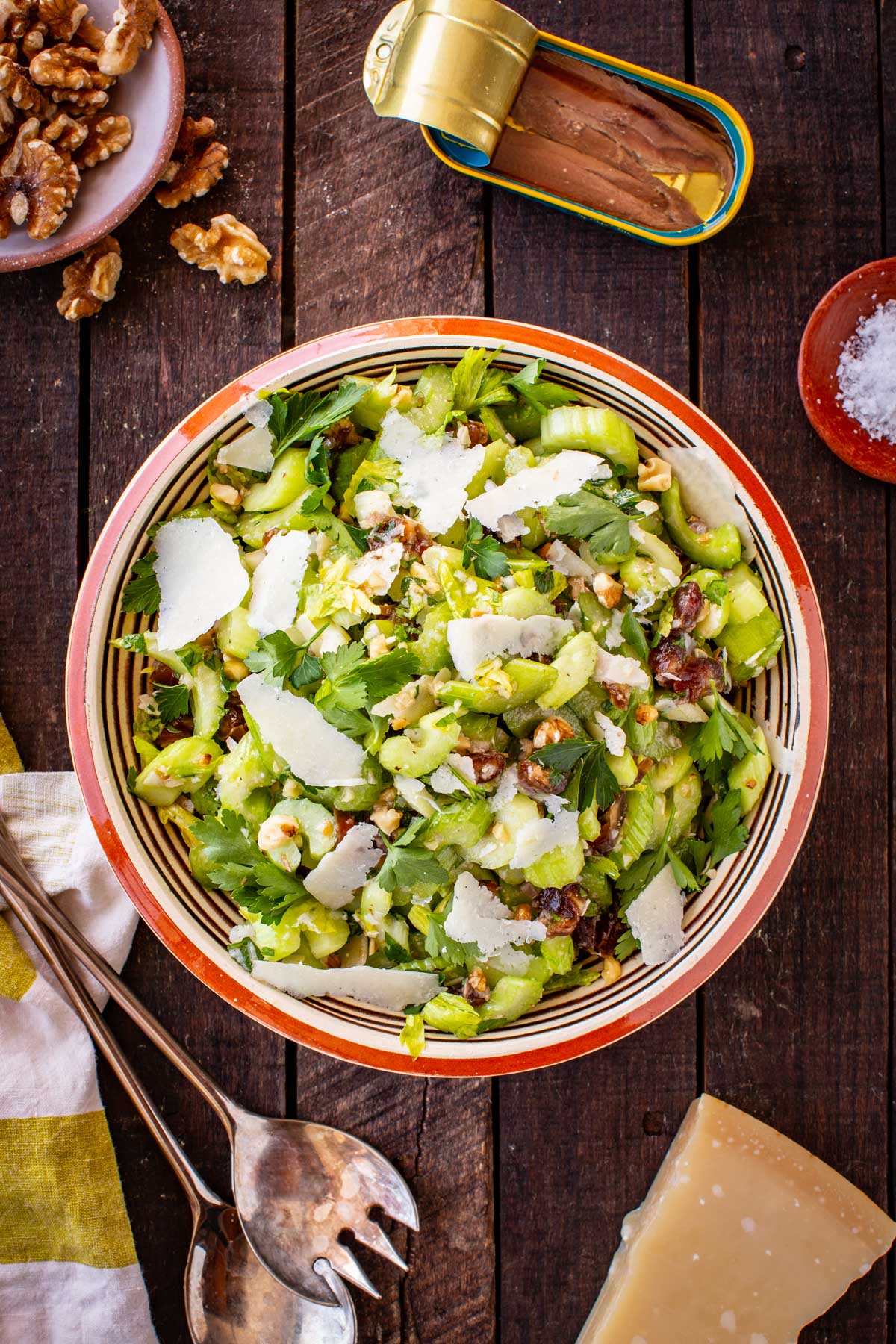 Full overhead shot of the celery salad in a striped ceramic bowl on a dark wood surface, surrounded by a tin of anchovies, a wedge of Parmesan, a dish of walnuts, flaky salt, and serving utensils.