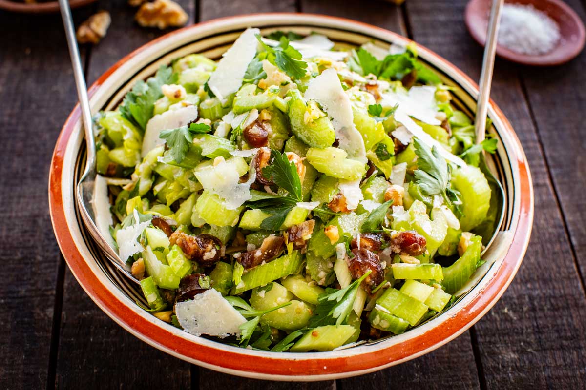 Angled close-up of the celery salad piled high in a striped ceramic bowl with two serving spoons, showing chunky celery pieces, date pieces, walnut bits, parsley, and generous Parmesan shavings.