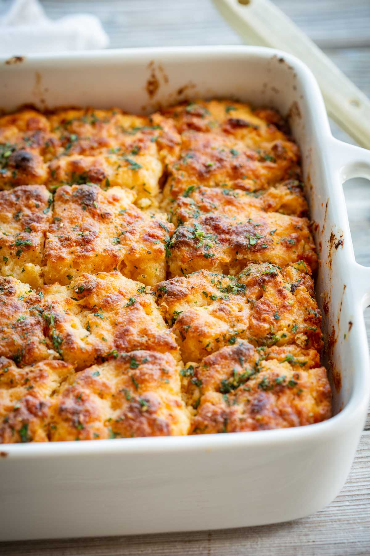 Golden brown cheddar bay butter swim biscuits scored into squares in a white ceramic baking dish, topped with chopped fresh parsley.