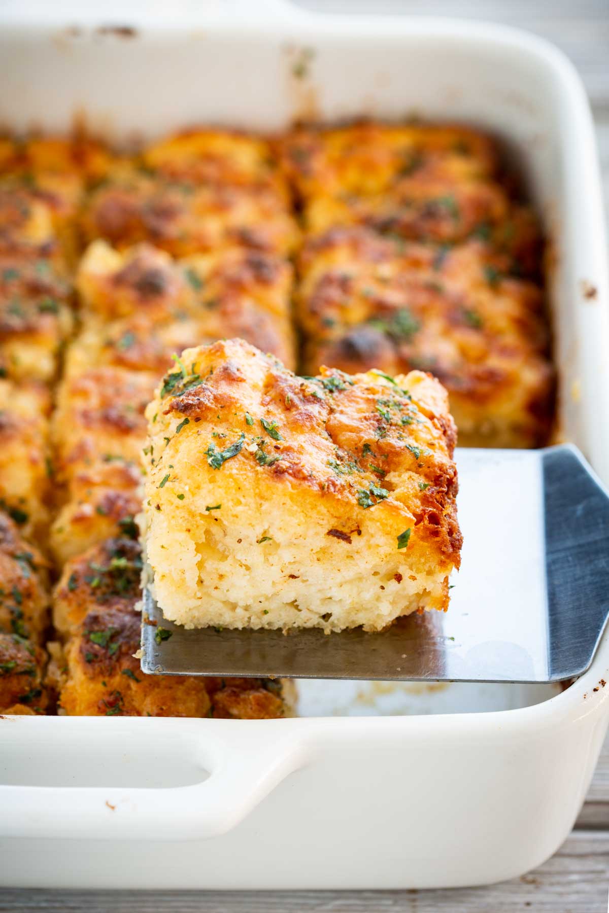 A single square of cheddar bay butter swim biscuit being lifted from a white baking dish with a metal spatula, showing a fluffy, tender interior.