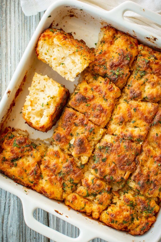 Overhead view of cheddar bay butter swim biscuits in a white baking dish, with one biscuit broken in half to reveal a soft, pillowy crumb.