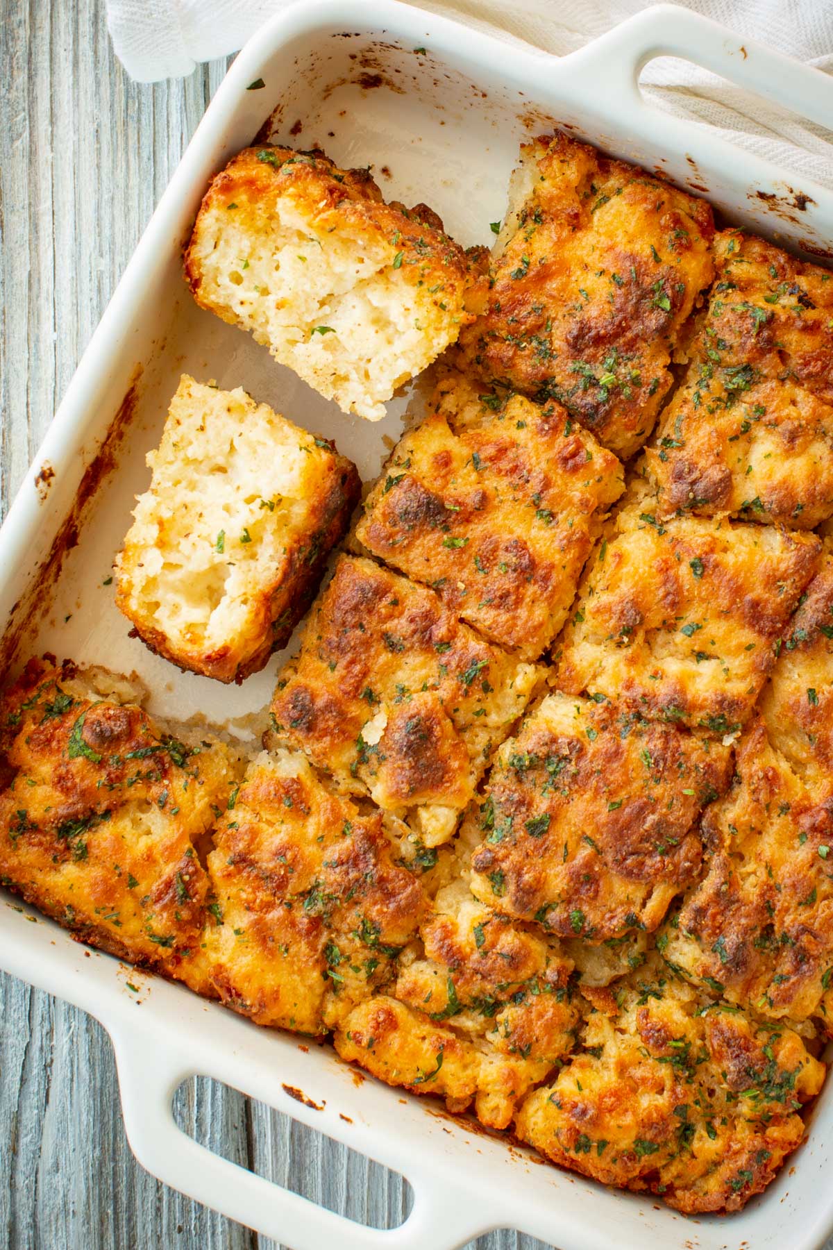 Overhead view of cheddar bay butter swim biscuits in a white baking dish, with one biscuit broken in half to reveal a soft, pillowy crumb.