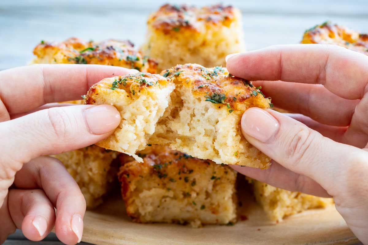 Two hands pulling apart a cheddar bay butter swim biscuit to reveal a soft, cheesy interior, with more biscuits stacked on a wooden board in the background.